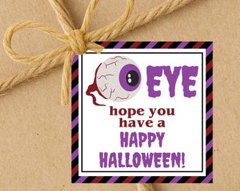 May include: A Halloween-themed gift tag with a cartoon eyeball graphic and the word "EYE" in purple. The tag also includes the message "hope you have a HAPPY HALLOWEEN!" in purple text, with a black, white, and purple border. The tag is tied with twine.