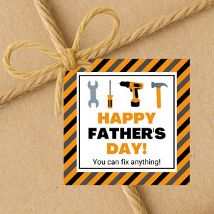 Puede incluir: Una etiqueta de regalo con un lazo de cuerda marrón. La etiqueta tiene un fondo blanco con rayas diagonales naranjas y negras. Presenta ilustraciones de herramientas y el texto "HAPPY FATHER'S DAY! You can fix anything!"