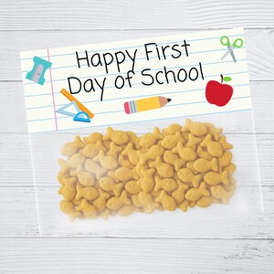 May include: A clear plastic bag filled with goldfish crackers. The bag has a label with the text "Happy First Day of School" and school-themed illustrations like a pencil, apple, and scissors.