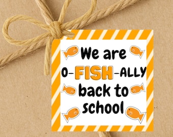 Op de afbeelding: Een vierkant cadeaulabel met de tekst "We are o-FISH-ally back to school" in zwart en oranje. Het label heeft oranje en witte diagonale strepen en kleine oranje visillustraties. Het label is met touw aan een bruin pakket bevestigd.