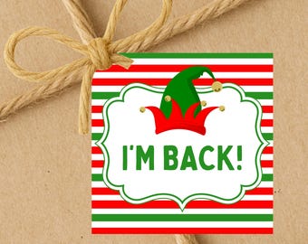 May include: A festive gift tag with red and green stripes and the words "I'M BACK!". The tag features a green elf hat with a red crown and gold bells. The tag is tied with a brown twine bow.