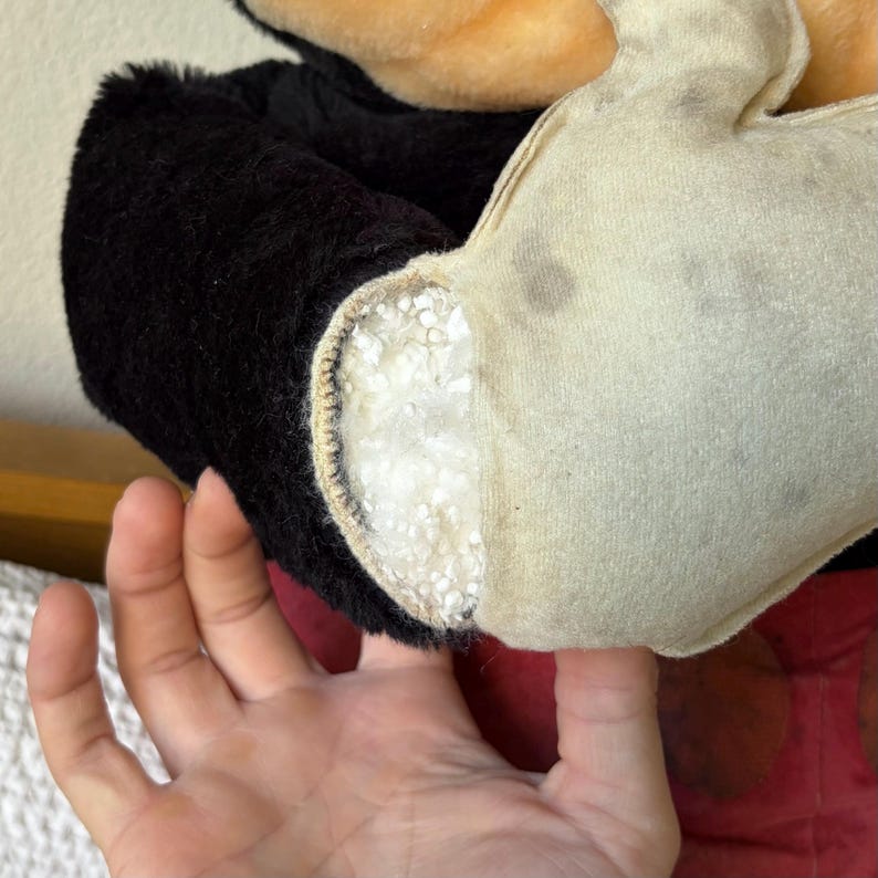 May include: Close-up of a vintage Mickey Mouse plush toy. The black plush arm and white glove are visible, with exposed stuffing. The red shorts are partially visible. The toy shows signs of wear and tear.