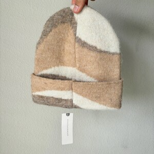 May include: A knit beanie in a geometric pattern of beige, cream, and brown. The folded brim and the body of the hat feature a textured, soft material. A white tag with black text is attached to the hat.