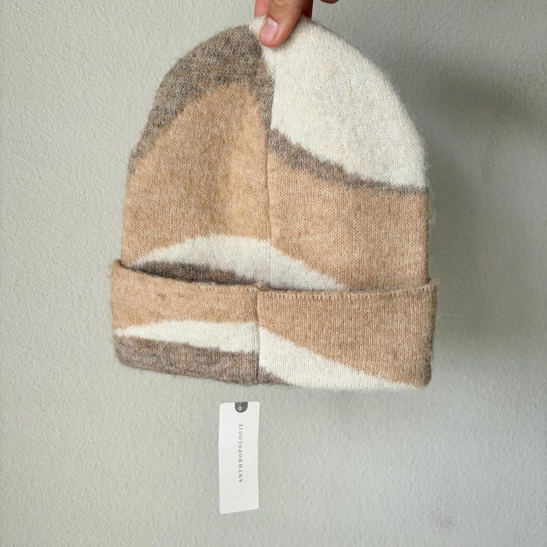 May include: A knit beanie in a geometric pattern of beige, cream, and brown. The folded brim and the body of the hat feature a textured, soft material. A white tag with black text is attached to the hat.