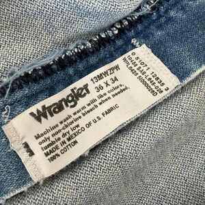 May include: Close-up of a light blue denim fabric with a white label. The label reads "Wrangler" and includes size information: 36 x 34 inches. The label also provides washing instructions and fabric composition: 100% cotton.