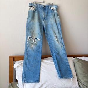 May include: Light blue denim jeans with distressed details, including rips at the knees and pockets. The jeans are hanging on a metal hanger, showcasing a vintage, worn aesthetic. The jeans have a classic five-pocket design.