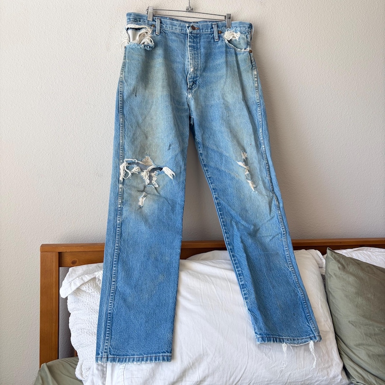 May include: Light blue denim jeans with distressed details, including rips at the knees and pockets. The jeans are hanging on a metal hanger, showcasing a vintage, worn aesthetic. The jeans have a classic five-pocket design.