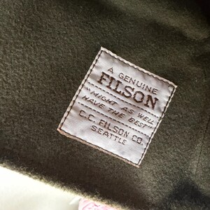 May include: A square white fabric label stitched onto olive green felted wool material. The label features dark brown text reading A GENUINE FILSON, MIGHT AS WELL HAVE THE BEST, and C.C. FILSON CO. SEATTLE, with a horizontal line separating the text.