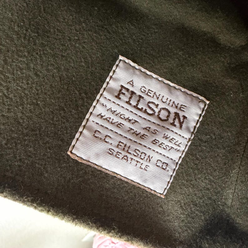 May include: A square white fabric label stitched onto olive green felted wool material. The label features dark brown text reading A GENUINE FILSON, MIGHT AS WELL HAVE THE BEST, and C.C. FILSON CO. SEATTLE, with a horizontal line separating the text.