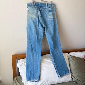 May include: Light blue denim jeans with a classic five-pocket design. The jeans feature a faded wash and the Wrangler logo on the back pockets. They are hanging on a metal hanger.