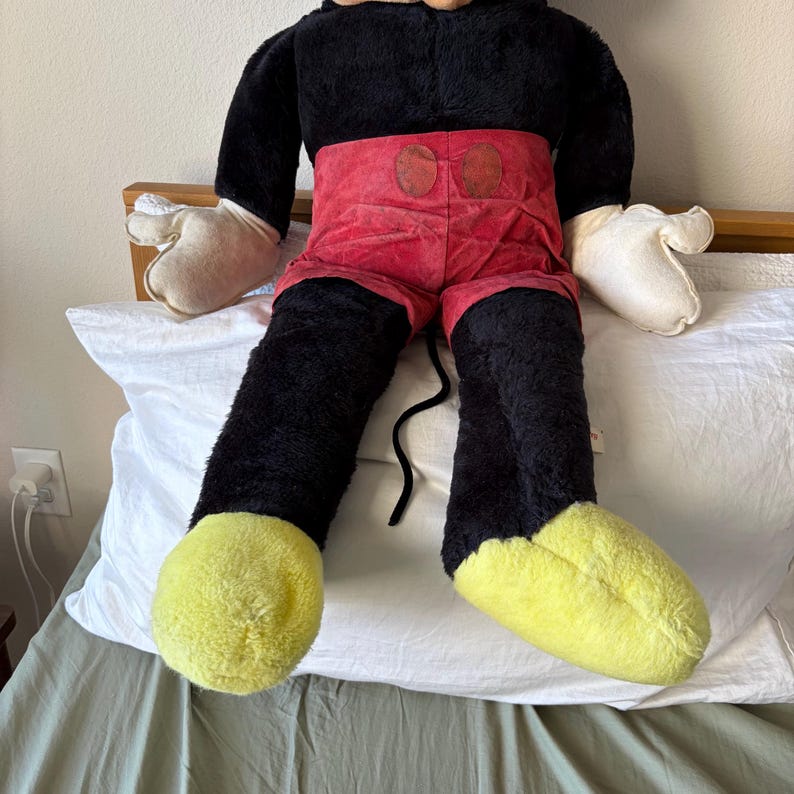May include: A large plush Mickey Mouse doll. The doll has a black body, red shorts, yellow feet, and white gloves. The doll is sitting on a white pillow.