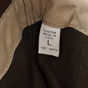 May include: A tan fabric interior lining with a white rectangular garment tag featuring black text that reads CUSTOM MADE IN U.S.A. L LOT #307U. The tag is stitched into the seam of a dark brown fabric garment with visible tan stitching.