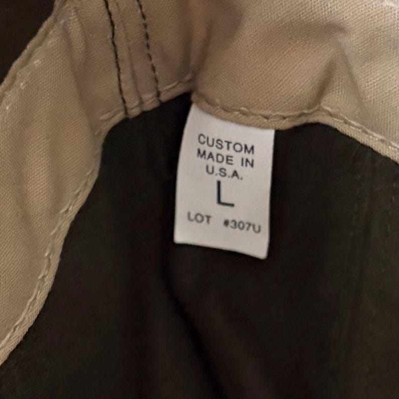 May include: A tan fabric interior lining with a white rectangular garment tag featuring black text that reads CUSTOM MADE IN U.S.A. L LOT #307U. The tag is stitched into the seam of a dark brown fabric garment with visible tan stitching.