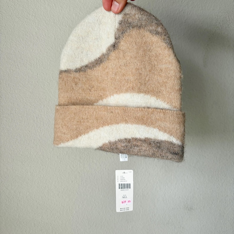 May include: A knit beanie in shades of beige, brown, and cream, with a folded brim. The hat features a wavy pattern. A white tag with a barcode and text hangs from the hat.