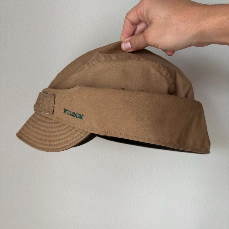May include: A tan-colored cap with a short, curved brim and a wide, wrap-around band. The word FILSON is embroidered in dark green thread on the side of the band. The cap features visible stitching along the seams and a structured crown design.