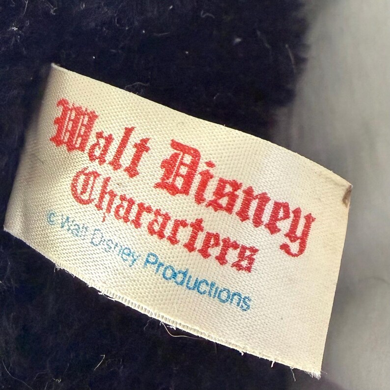 May include: A close-up of a white tag with red text that reads "Walt Disney Characters". Below the text is "&copy; Walt Disney Productions" in blue. The tag is attached to a dark blue plush toy.