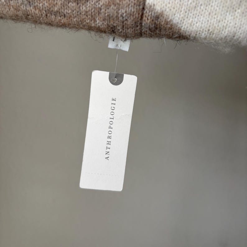 May include: A white tag with the word "ANTHROPOLOGIE" printed in black. The tag is attached to a brown and white patterned fabric. The background is a solid, neutral color.
