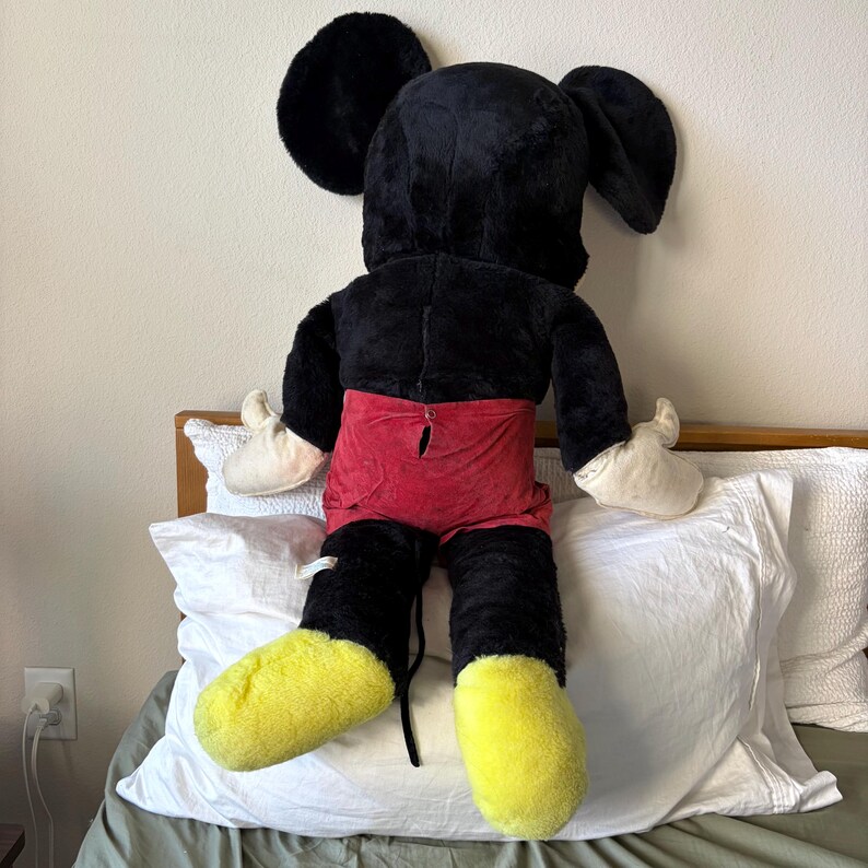 May include: A large plush Mickey Mouse doll, viewed from the back. The doll features a black body and head with large ears, red shorts, white gloves, and yellow feet. The doll is sitting on a bed with white pillows.