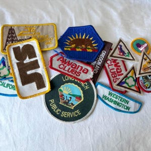 May include: A collection of embroidered patches in various shapes and sizes. The patches feature text and graphics, including a bear, a lighthouse, and the words "Awana Clubs" and "Western Washington". Colors include blue, green, brown, and white.
