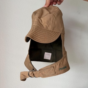 May include: A tan canvas cap with a curved brim and an extended neck flap. The cap features a dark green interior lining, a white rectangular label with the text FILSON, and a metal D-ring closure on the side with the word FILSON embroidered in green.
