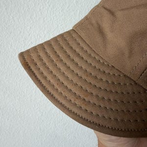 May include: A tan bucket hat made of a durable, woven fabric. The brim features multiple rows of parallel, decorative stitching in a matching thread color, providing structure and detail to the circular design of the hat's lower section.