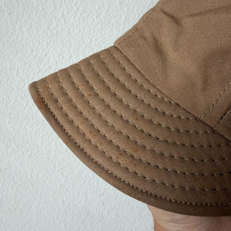 May include: A tan bucket hat made of a durable, woven fabric. The brim features multiple rows of parallel, decorative stitching in a matching thread color, providing structure and detail to the circular design of the hat's lower section.