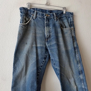 May include: Faded blue denim jeans hanging on a metal hanger. The jeans have a classic five-pocket design, with visible wear and tear, including distressed edges on the front pockets and light discoloration. The jeans are a medium wash.