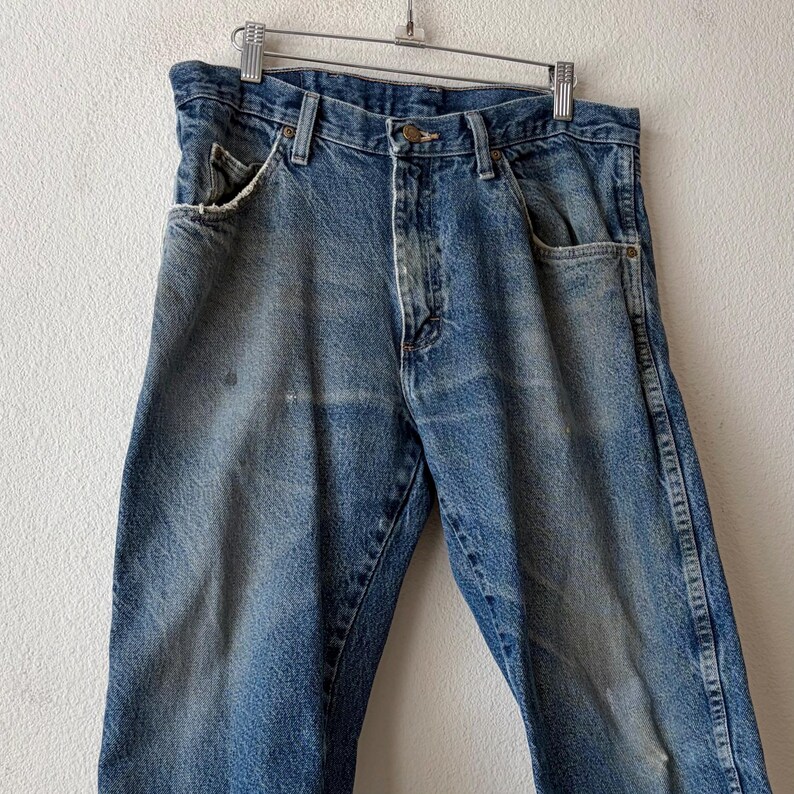 May include: Faded blue denim jeans hanging on a metal hanger. The jeans have a classic five-pocket design, with visible wear and tear, including distressed edges on the front pockets and light discoloration. The jeans are a medium wash.