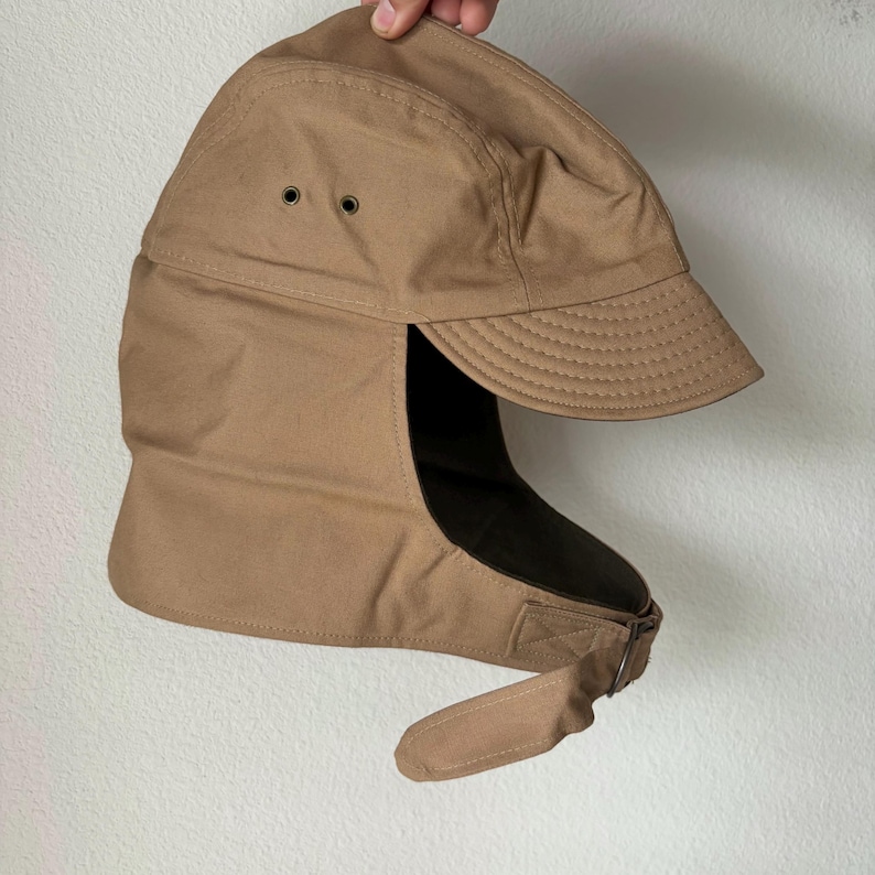 May include: A tan canvas cap with a short brim and ear flaps that extend downward. The cap features two small metal ventilation eyelets on the side and a fabric strap with a metal buckle closure attached to the lower edge of the ear flap section.