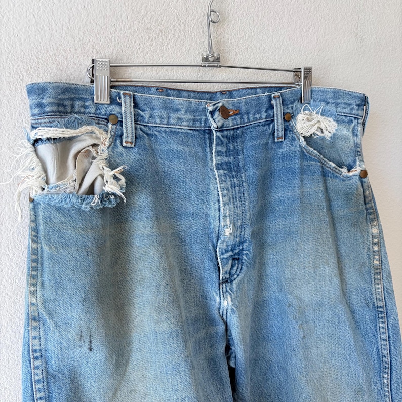 May include: Light blue denim jeans with distressed details, including ripped pockets and faded areas. The jeans are hanging on a metal hanger against a white background. The jeans have a button closure and classic five-pocket design.