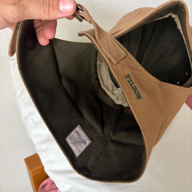 May include: A tan cap with a dark olive green fleece lining and ear flaps. The exterior features the word FILSON embroidered in dark green thread. A white fabric label inside reads A GENUINE FILSON GARMENT, C.C. FILSON CO. SEATTLE.