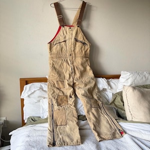 May include: Tan canvas dungarees featuring brown adjustable shoulder straps, a red interior lining, and multiple zipped pockets on the chest. The legs include several stitched-on patches and full-length side zips extending from the ankles.