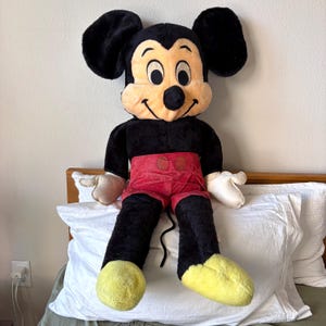 May include: A large plush Mickey Mouse doll. The doll has a black body, large black ears, and a tan face with a smiling expression. It wears red shorts, white gloves, and yellow shoes. The doll is sitting on a white pillow.