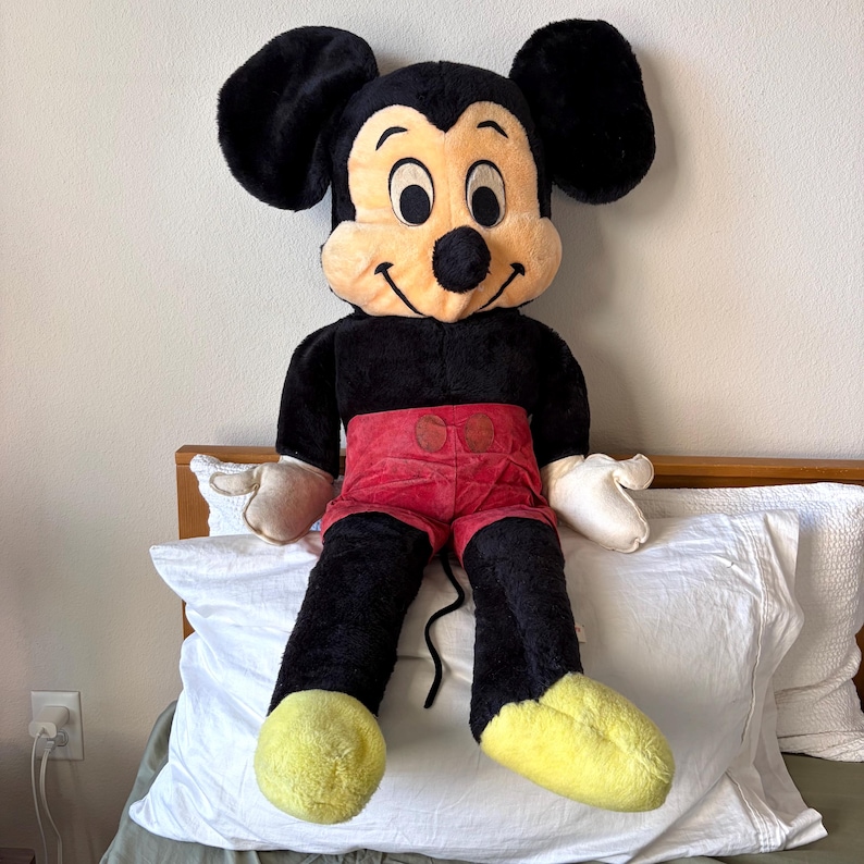 May include: A large plush Mickey Mouse doll. The doll has a black body, large black ears, and a tan face with a smiling expression. It wears red shorts, white gloves, and yellow shoes. The doll is sitting on a white pillow.