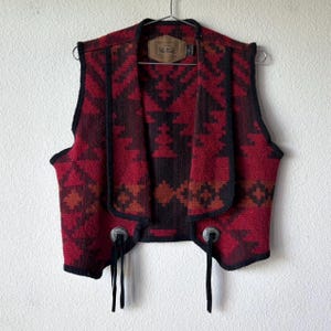 Puede incluir: Un chaleco estampado en rojo y negro con un diseño del suroeste. El chaleco tiene un cuello chal y ribetes negros. Cuenta con detalles metálicos plateados y lazos negros en la parte delantera.