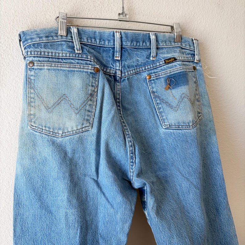 May include: Light blue denim jeans with the Wrangler logo on the back pocket. The jeans are hanging on a metal hanger. The jeans have a classic five-pocket design and a faded wash.