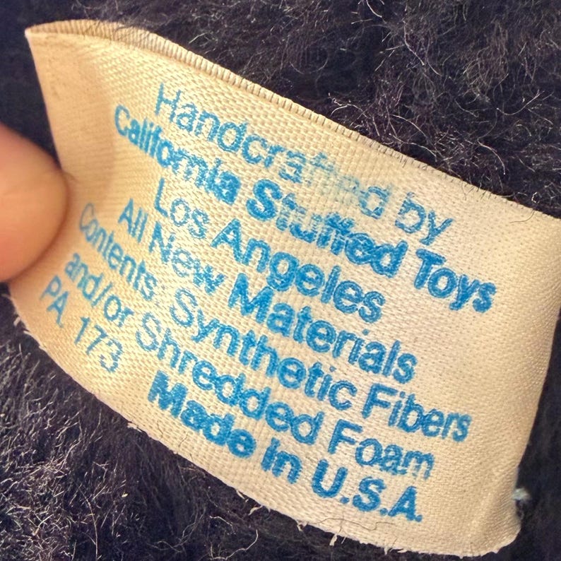 May include: A close-up of a tag from a stuffed toy. The tag is cream-colored with blue text that reads "Handcrafted by California Stuffed Toys, Los Angeles, All New Materials, Contents: Synthetic Fibers and/or Shredded Foam, Made in U.S.A."