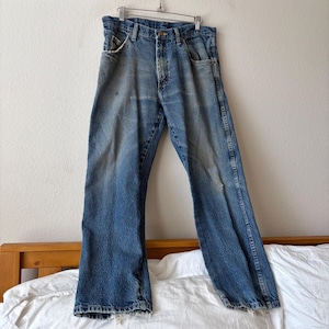 May include: Faded blue denim jeans hanging on a metal hanger. The jeans have a classic five-pocket design, a button and zipper closure, and show signs of wear and fading. The jeans are displayed against a neutral background.