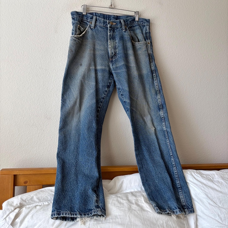 May include: Faded blue denim jeans hanging on a metal hanger. The jeans have a classic five-pocket design, a button and zipper closure, and show signs of wear and fading. The jeans are displayed against a neutral background.