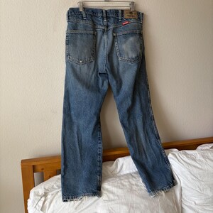 May include: Faded blue denim jeans hanging on a metal hanger. The jeans have a classic five-pocket design, with worn patches on the back pockets and knees. A red and white label is visible on the back.