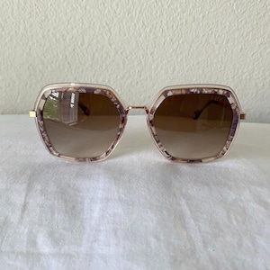 May include: Hexagonal-shaped sunglasses with a light brown gradient lens. The frames are a light gold color with a decorative pattern in shades of purple and brown. The sunglasses have a modern, stylish design.
