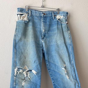 May include: Light blue denim jeans with distressed details, including rips and frayed edges. The jeans are hanging on a metal hanger. The jeans have a button and zipper closure.