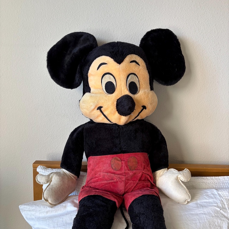 May include: A large plush Mickey Mouse doll. The doll has a black body, large black ears, a tan face, and red shorts. The doll's hands are white, and it is sitting on a bed with a white sheet.