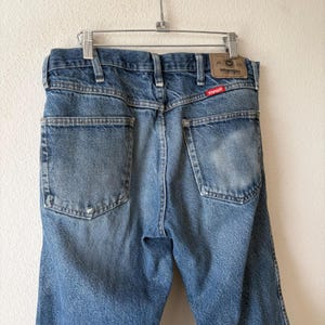 May include: Light blue denim jeans hanging on a metal hanger. The jeans have two back pockets and a Wrangler brand patch. The Wrangler logo is visible on the back pocket. The jeans are faded and worn.