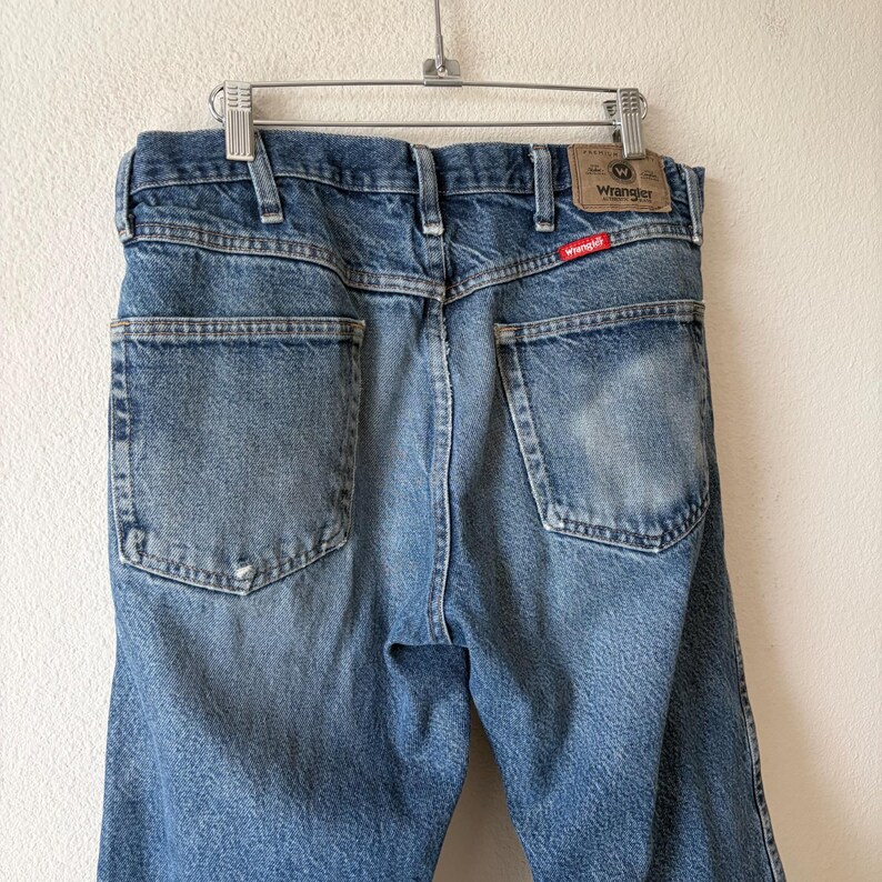 May include: Light blue denim jeans hanging on a metal hanger. The jeans have two back pockets and a Wrangler brand patch. The Wrangler logo is visible on the back pocket. The jeans are faded and worn.
