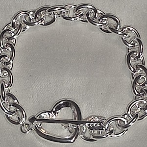 May include: A silver-toned bracelet featuring a chain link design. The bracelet has a heart-shaped clasp with an arrow through it. The jewelry is likely made of sterling silver or a similar metal. The bracelet is a piece of fashion jewelry.