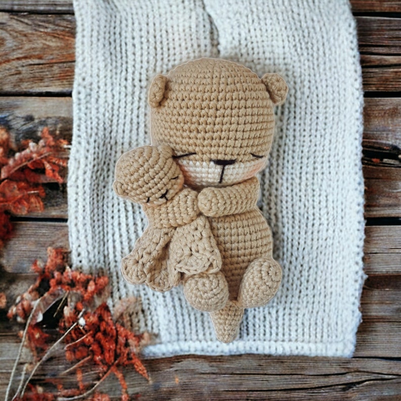 May include: A light brown crocheted teddy bear embracing a smaller bear, both lying on a white knitted blanket. The background features a wooden surface. The bear has closed eyes and a small black nose.