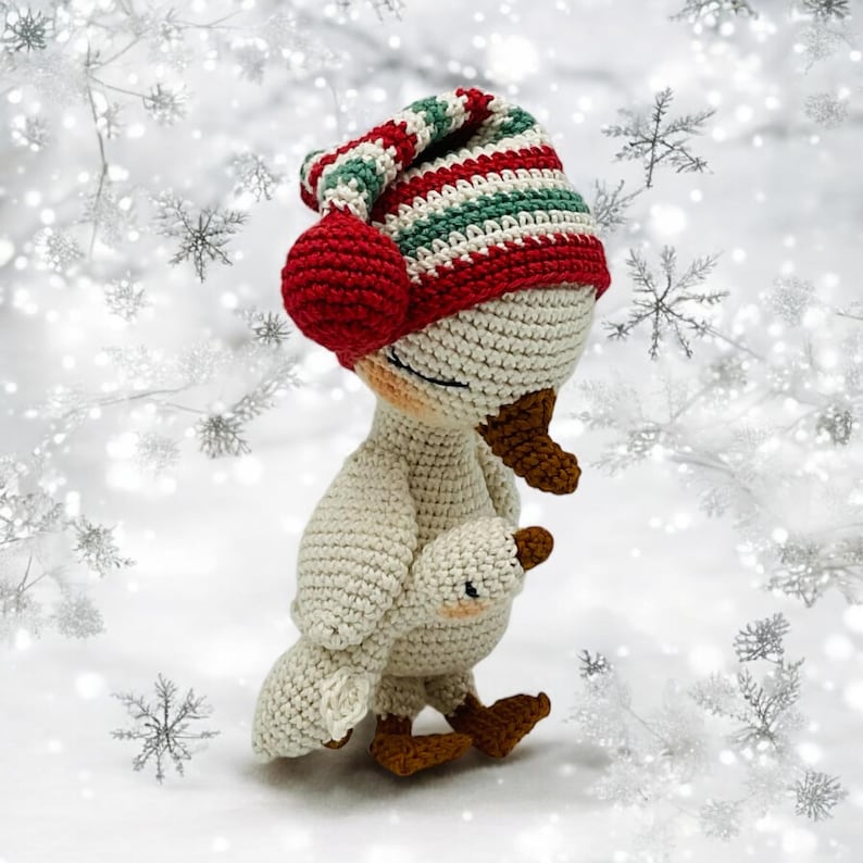 May include: A crocheted goose wearing a red, green, and white striped hat with a red pom-pom. The goose is holding a smaller goose, both in cream-colored yarn. The background is a snowy scene with snowflakes.
