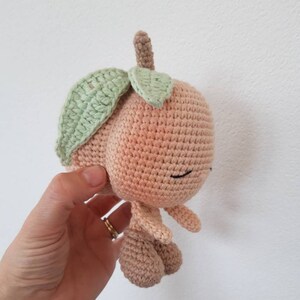 May include: A crocheted peach-colored plush toy with a green crocheted leaf on top. The toy has a smiling face with black eyes and a small mouth.
