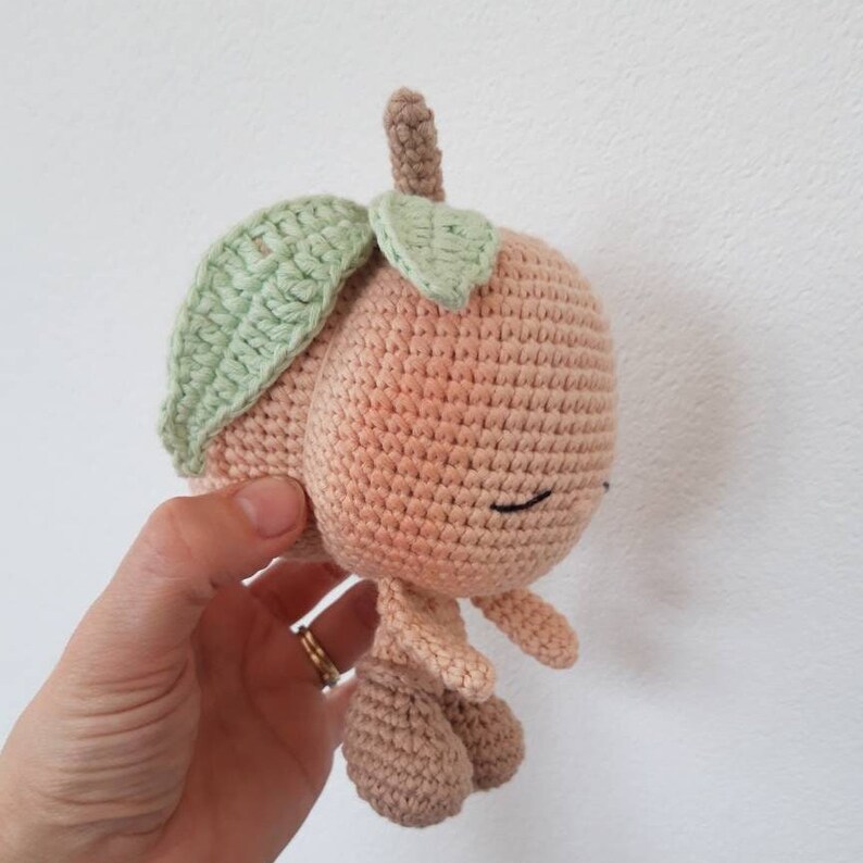 May include: A crocheted peach-colored plush toy with a green crocheted leaf on top. The toy has a smiling face with black eyes and a small mouth.